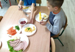 Jeden z chłopców sięga po talerzyk z jajecznicą, drugi chłopiec trzyma widelec i zastanawia się, co nałożyć sobie jeszcze na kanapkę